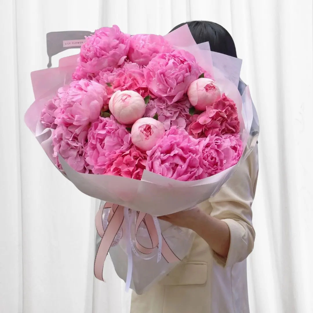 Blush Petals Peony Bouquet wrapped in white tissue paper with a pink satin ribbon, featuring 14 soft pink peonies in full bloom.