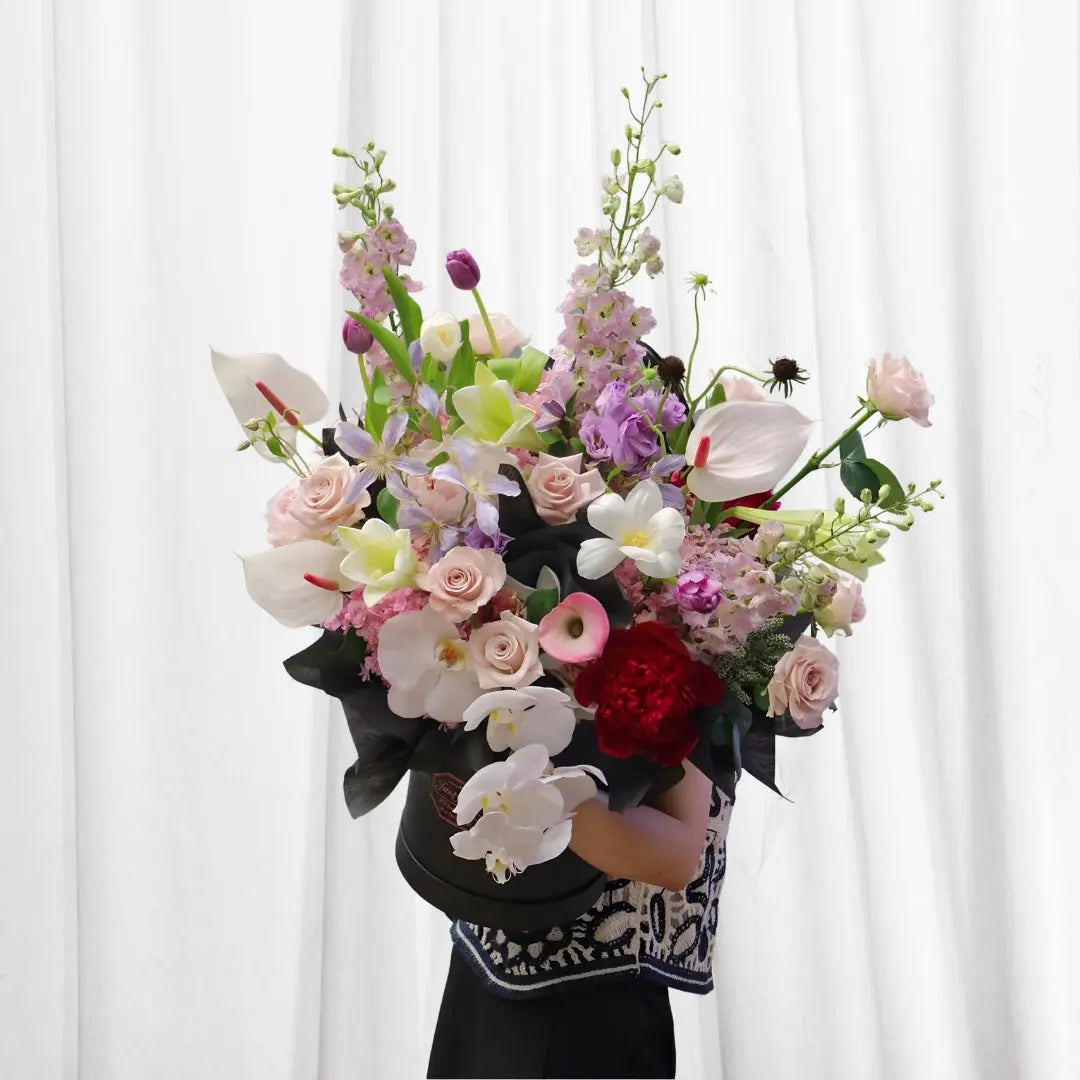 Blush Radiance Floral Symphony bouquet wrapped in red paper with a red ribbon, featuring pink roses, white calla lilies, orchids, peonies, delphinium, and tulips.