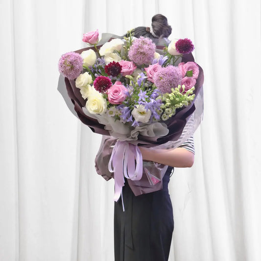 Elegant Romance Bouquet with pink roses, white roses, Allium, burgundy Gomphrena, purple Agapanthus, and green Lisianthus wrapped in purple and gray paper.