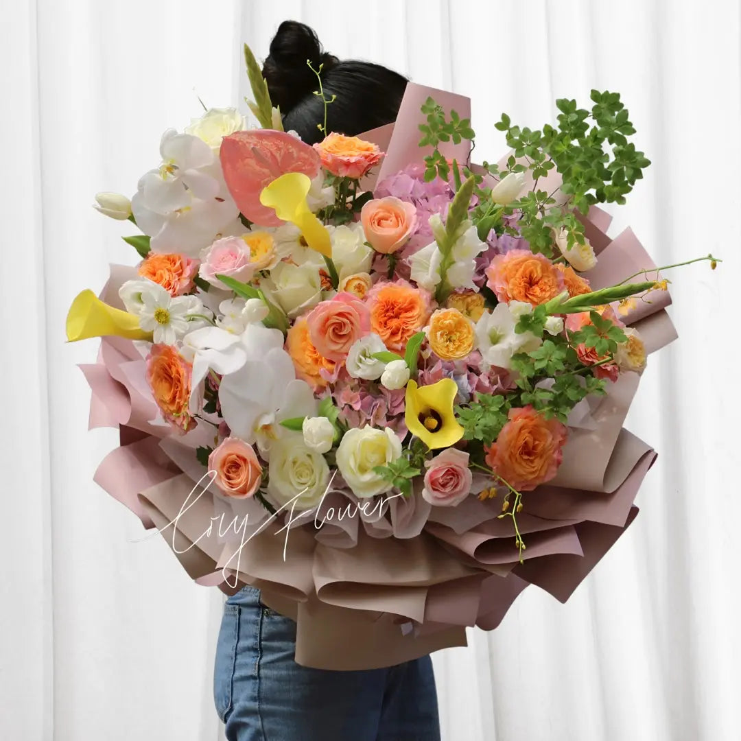 Golden Harmony Blossom Bouquet with peach roses, yellow calla lilies, white orchids, tulips, and hibiscus, wrapped in soft blush tissue paper.