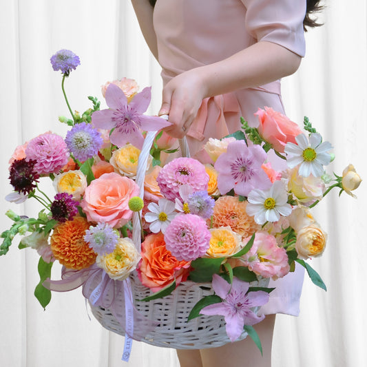 Serenity Blossom Garden bouquet in a white woven basket with pale pink satin ribbon, featuring peach roses, zinnias, lilies, cosmos, dahlias, and stock flowers.