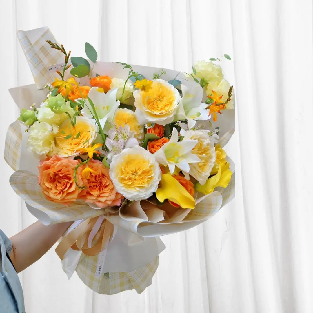 Sunny Day Blossom Bouquet featuring yellow roses, orange roses, white lilies, and calla lilies wrapped in yellow checkered tissue paper with a light brown ribbon.