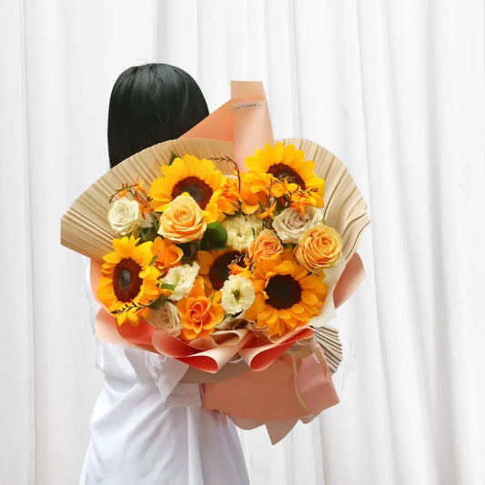 Sunset Glow Floral Bouquet with sunflowers, orange roses, peach roses, white lisianthus, freesia, and eucalyptus in peach wrap.