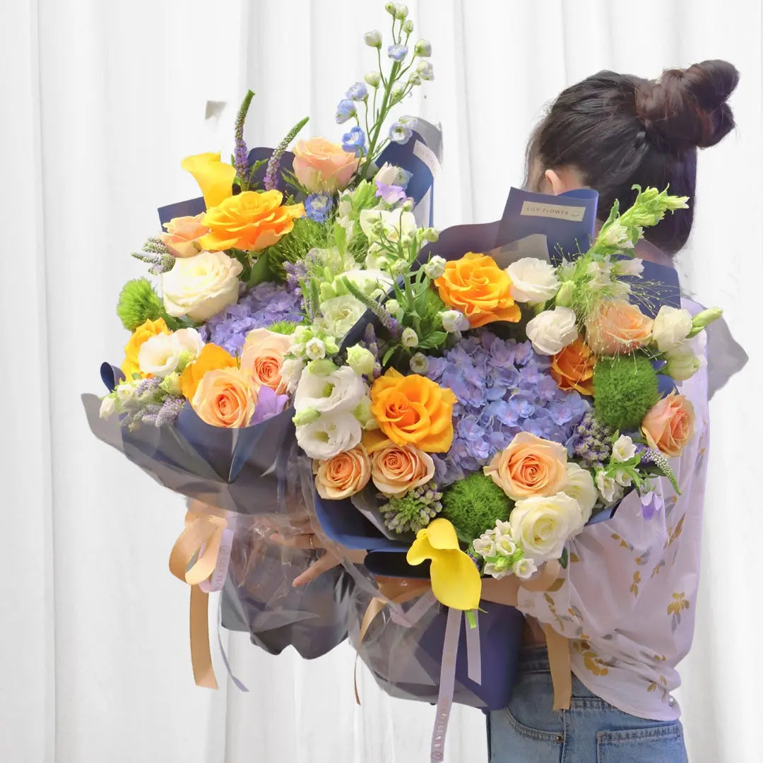 6 Orange Roses, 4 Peach Roses, 4 White Roses, 2 Blue Hydrangea Stems, 2 Yellow Calla Lilies, 2 Green Mums (Button Chrysanthemums), 6 Lisianthus Stems, Various Filler Flowers (Statice, Greenery)