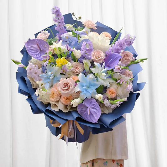 Vibrant Mystic Harmony Bouquet featuring soft peach roses, white lilies, blue lilies, purple anthuriums, and delphinium flowers