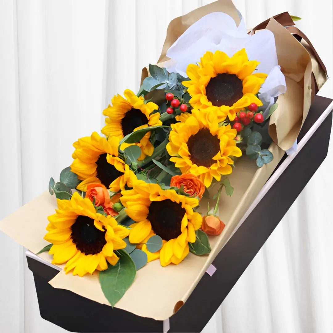 Vibrant Sunshine Floral Box featuring sunflowers, orange ranunculus, eucalyptus, and red berries in a sleek black floral box with a light pink ribbon